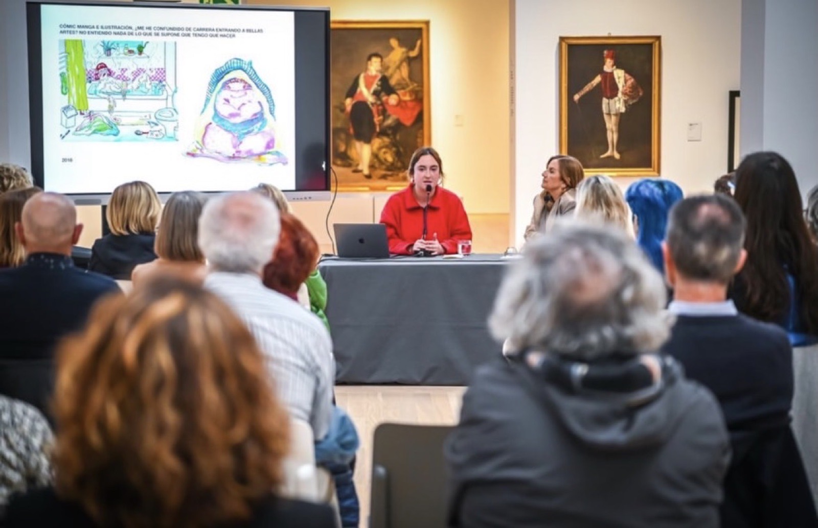 Una artista presenta su trabajo en una mesa ante el público, con dibujos proyectados en pantalla
